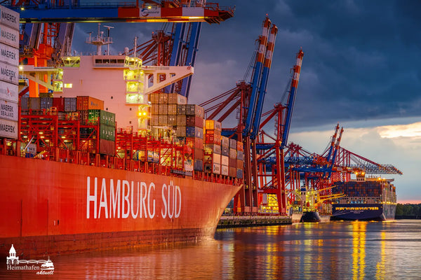 Das Containerschiff HAMBURG SÜD liegt am Waltershofer Hafen vor dramatischer Abendkulisse. Im Hintergrund ragen riesige Containerbrücken in den Himmel und weitere Containerriesen warten auf Abfertigung.