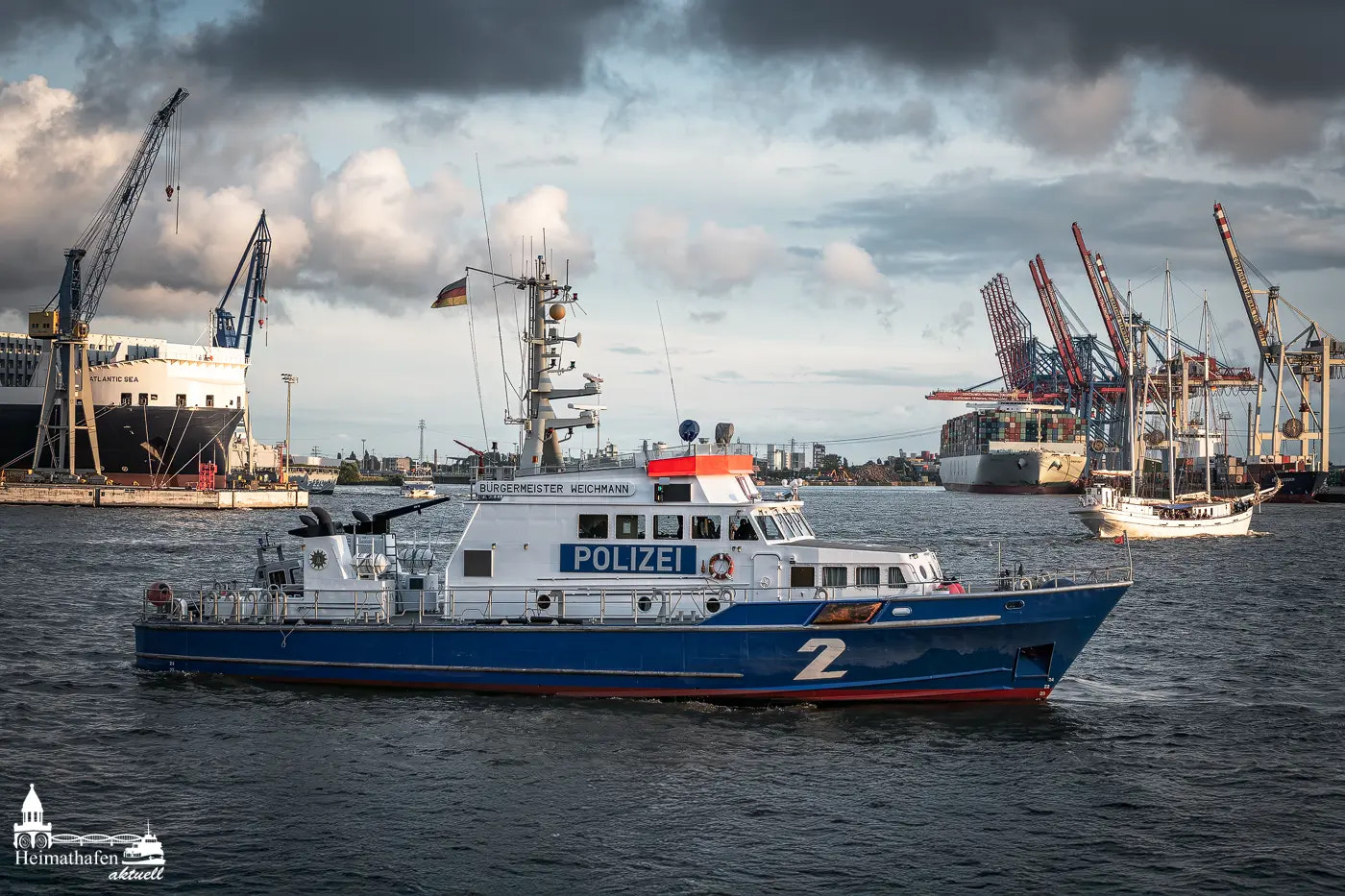 Polizeiboot „Bürgermeister Weichmann“ auf Streife im Hamburger Hafen mit Containerschiff und Kränen im Hintergrund.