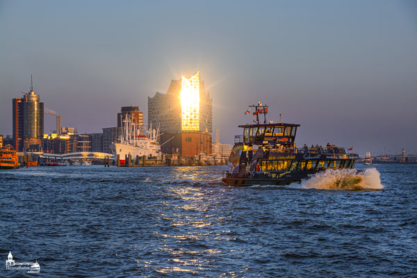 Lichterspiel auf der Elbe: Die Elbphilharmonie glänzt im Sonnenuntergang, während eine bunte Hafenfähre vorbeifährt.